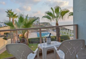 A balcony with two white plastic chairs, a small table, and a glass of champagne. Palm trees and a pool are visible in the background. - Sharon Hotel Herzliya (Herzliya)