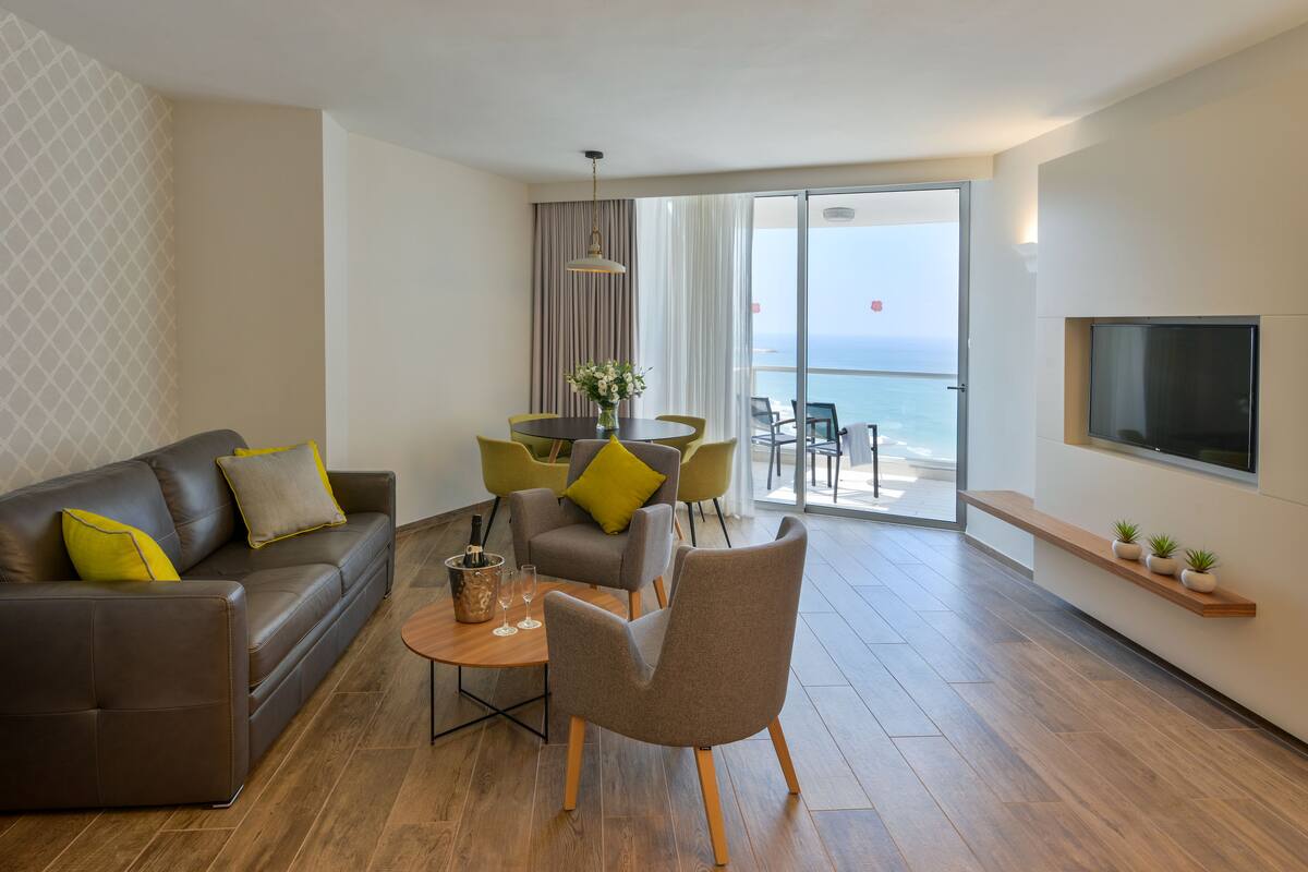 Un salon moderne avec un canapé en cuir, une table à manger avec des chaises, un téléviseur à écran plat fixé au mur et un balcon offrant une vue sur la mer.