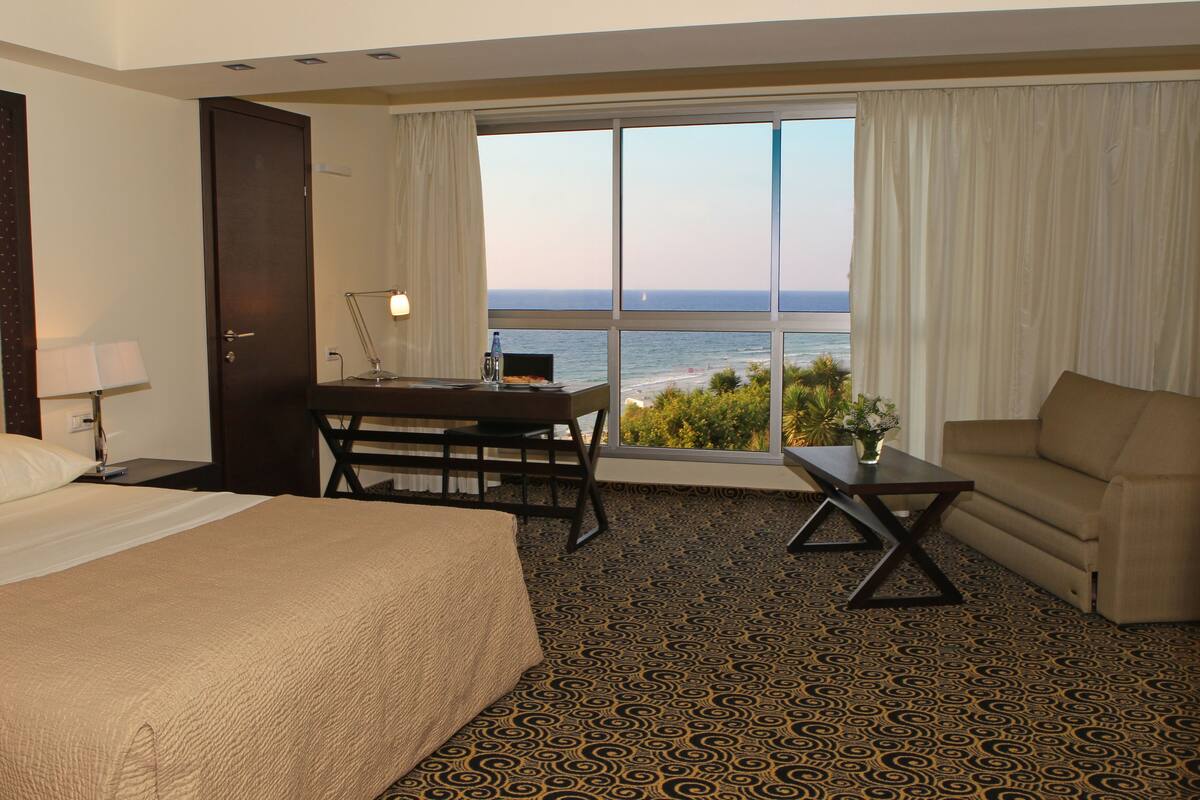 Une chambre d’hôtel comprenant un lit, un bureau, une chaise, un canapé et une grande fenêtre donnant sur la mer.