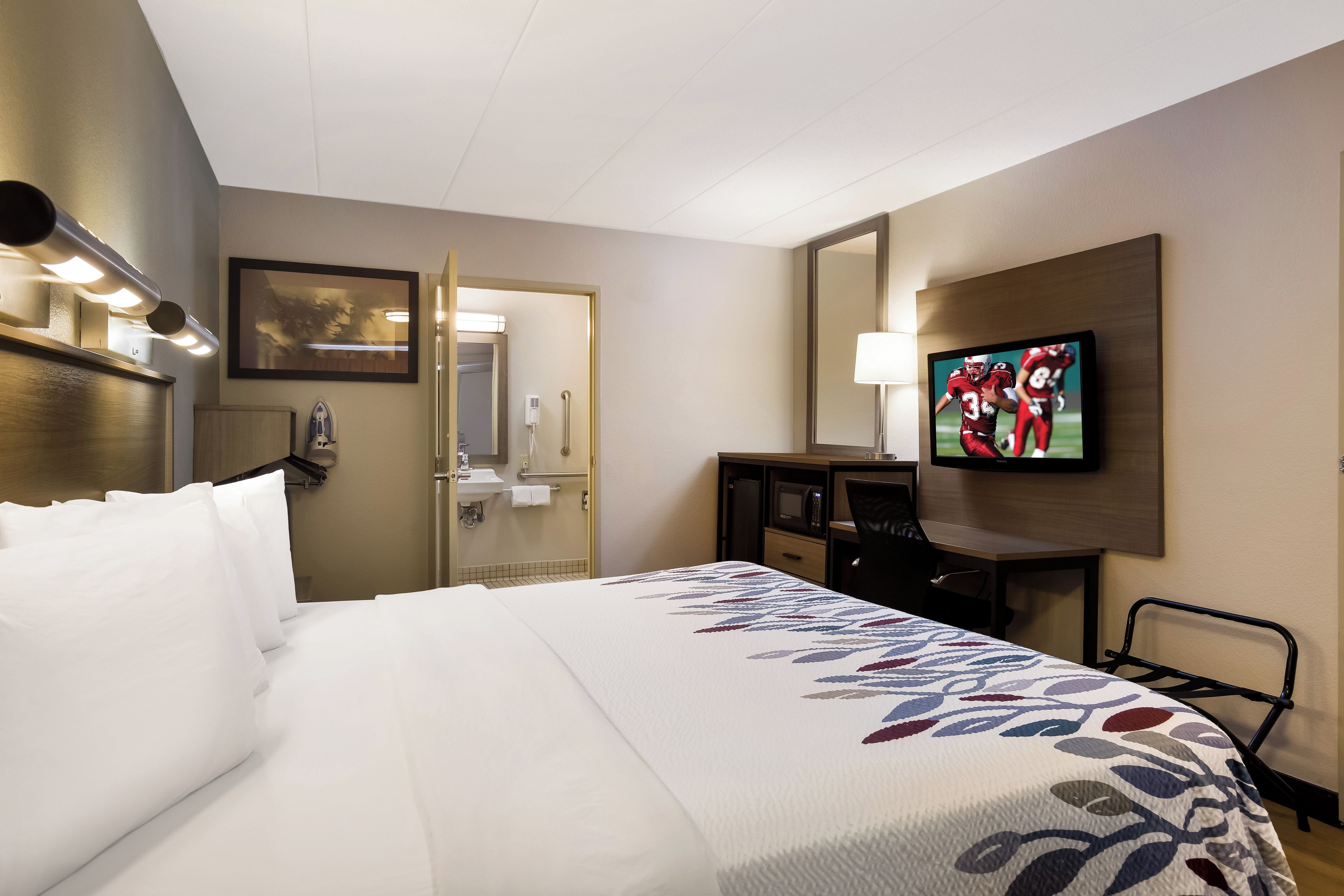 Quarto de hotel com cama, escrivaninha, televisão e banheiro visível por uma porta aberta.