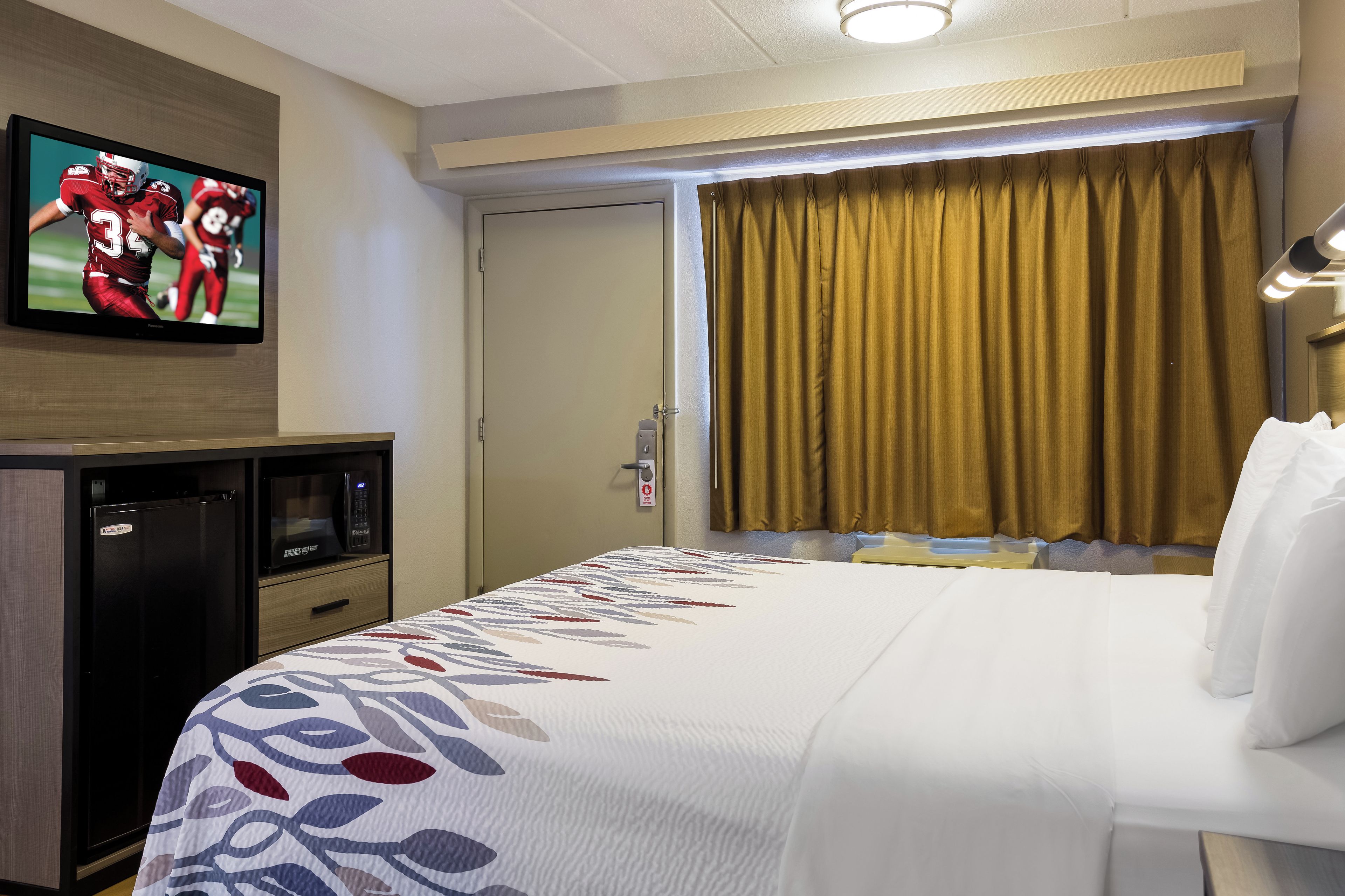 Een hotelkamer met een bed, een aan de muur bevestigde televisie, een magnetron en een raam met gordijnen.