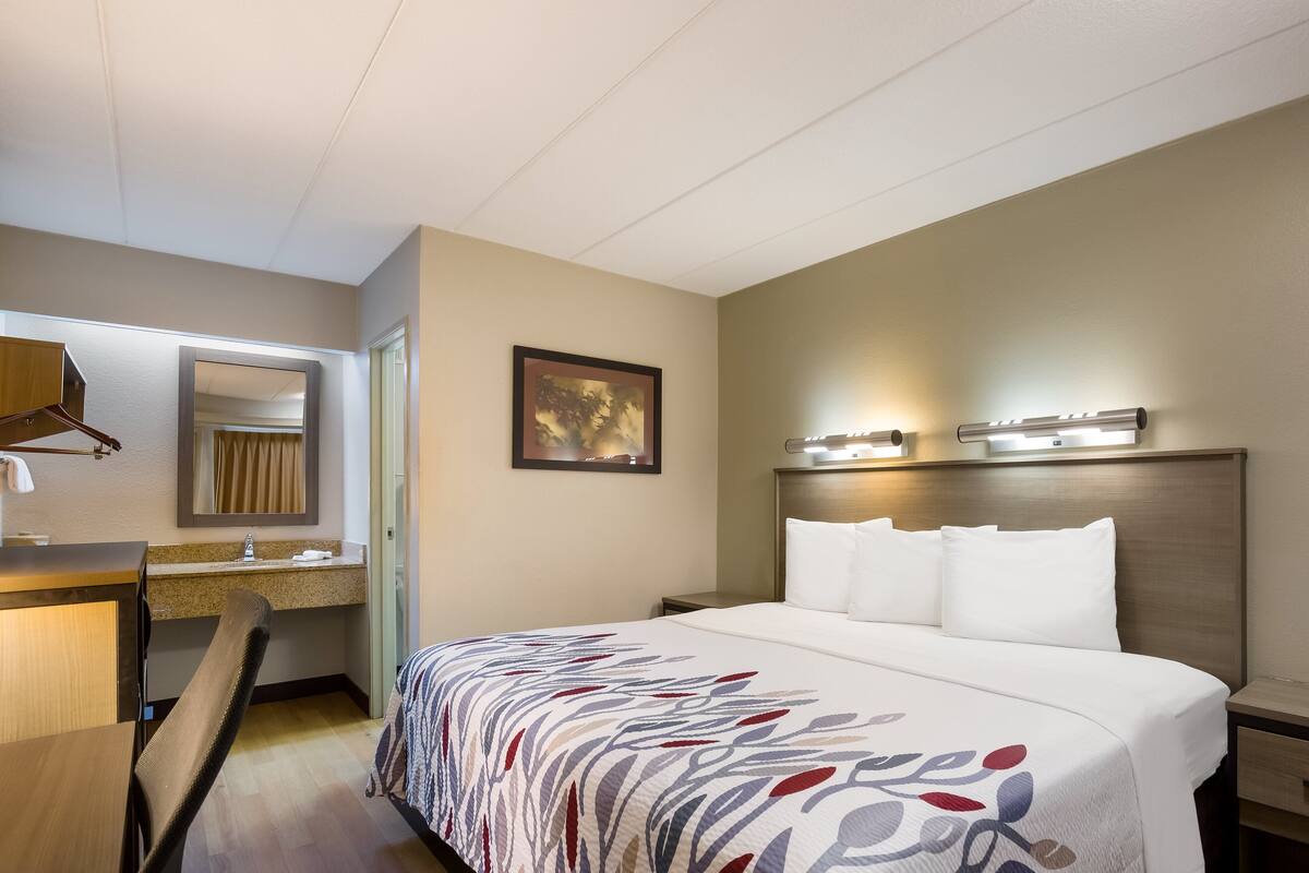 A hotel room with a bed, a desk, a chair, a mirror, a framed picture on the wall, and a nightstand with a lamp.