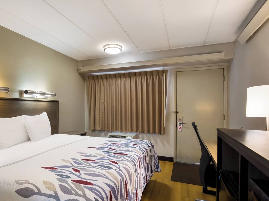 A hotel room with a bed, a desk, a lamp, a window with curtains, and a door.