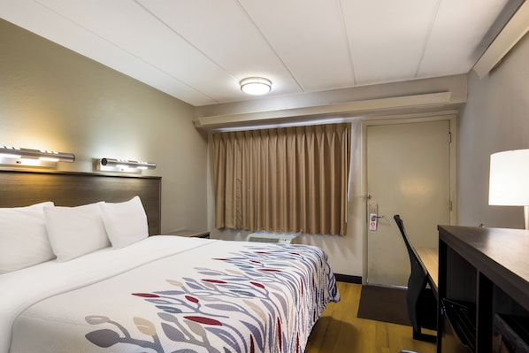 A hotel room with a bed, a desk, a lamp, a window with curtains, and a door.