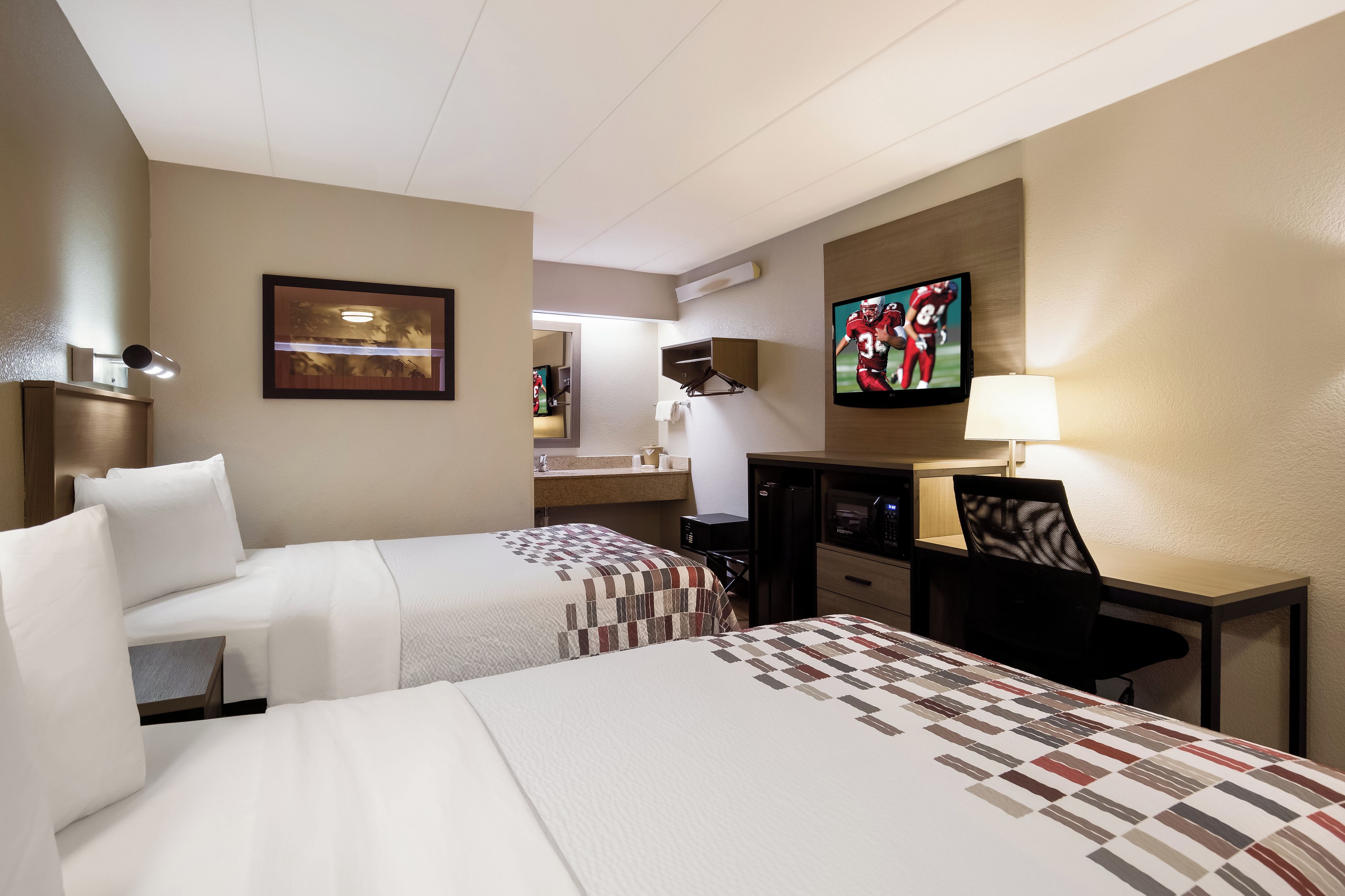 Habitación de hotel con dos camas, un escritorio, un televisor y un baño visible a través de una puerta abierta.