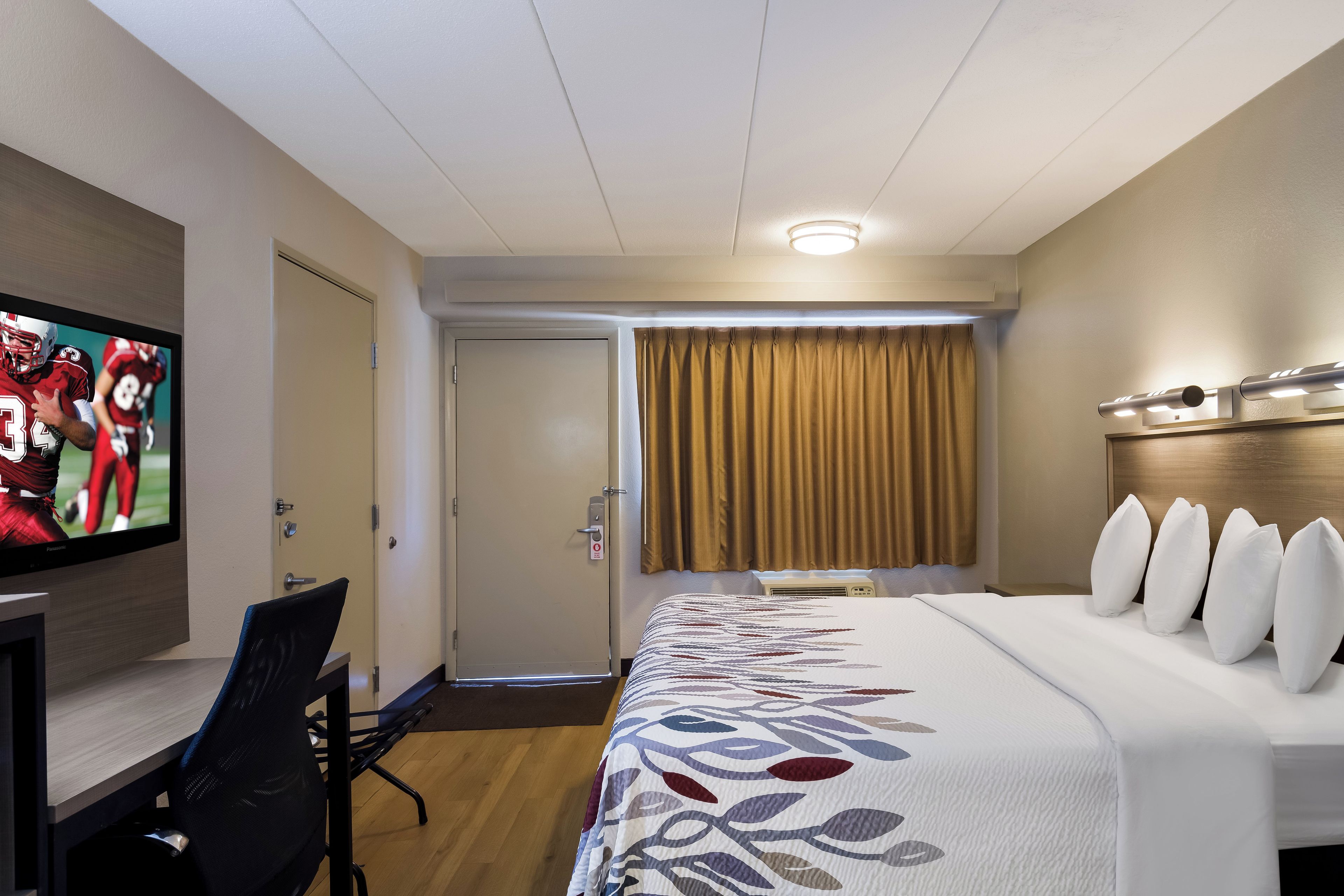 A hotel room with a bed, a TV, a desk, and a window with curtains.