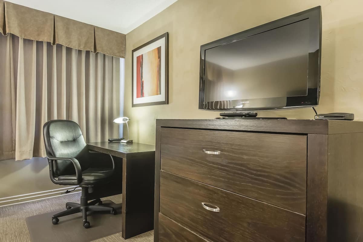 Habitación de hotel con un escritorio de madera oscura, una silla de oficina negra, un televisor de pantalla plana y una obra de arte enmarcada en la pared.