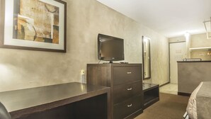 Una camera d'albergo con una scrivania in legno scuro, una televisione a schermo piatto, uno specchio, un letto e un angolo cottura.