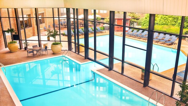 An indoor swimming pool with clear glass walls, lounge chairs, and potted plants. - Sheraton Harrisburg Hershey Hotel (Harrisburg)