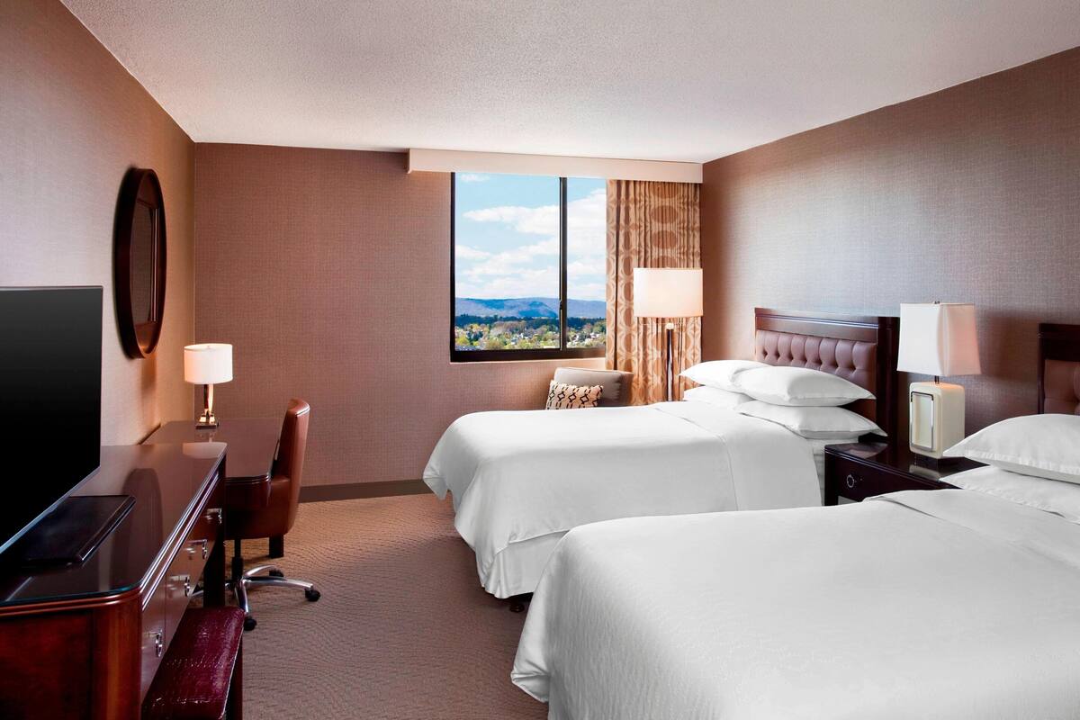 A hotel room with two beds, a desk, a chair, a TV, and a window with a mountain view.