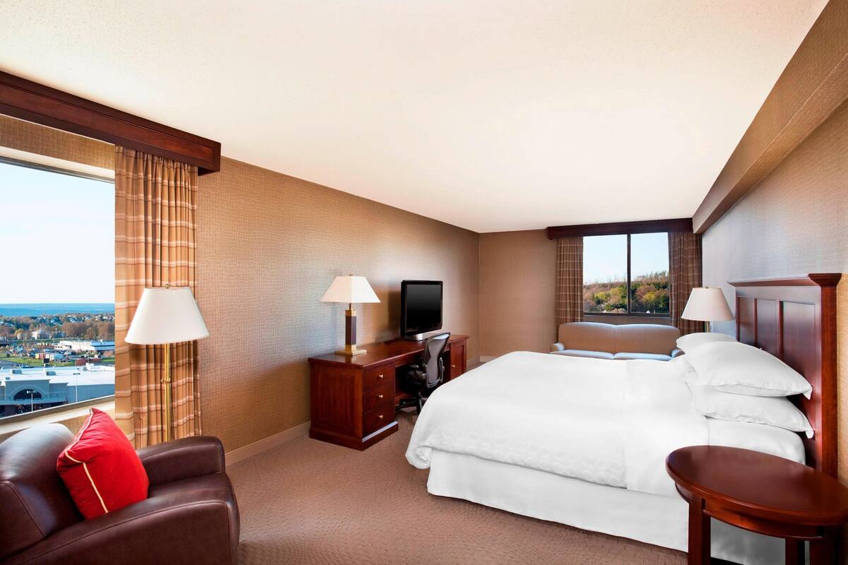 A hotel room with a bed, a desk with a computer, a TV, a chair, and a view of the city.