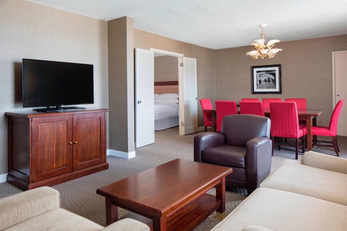 A hotel room with a flat-screen TV, a wooden cabinet, a leather armchair, a red dining table with chairs, a bed, and a chandelier.