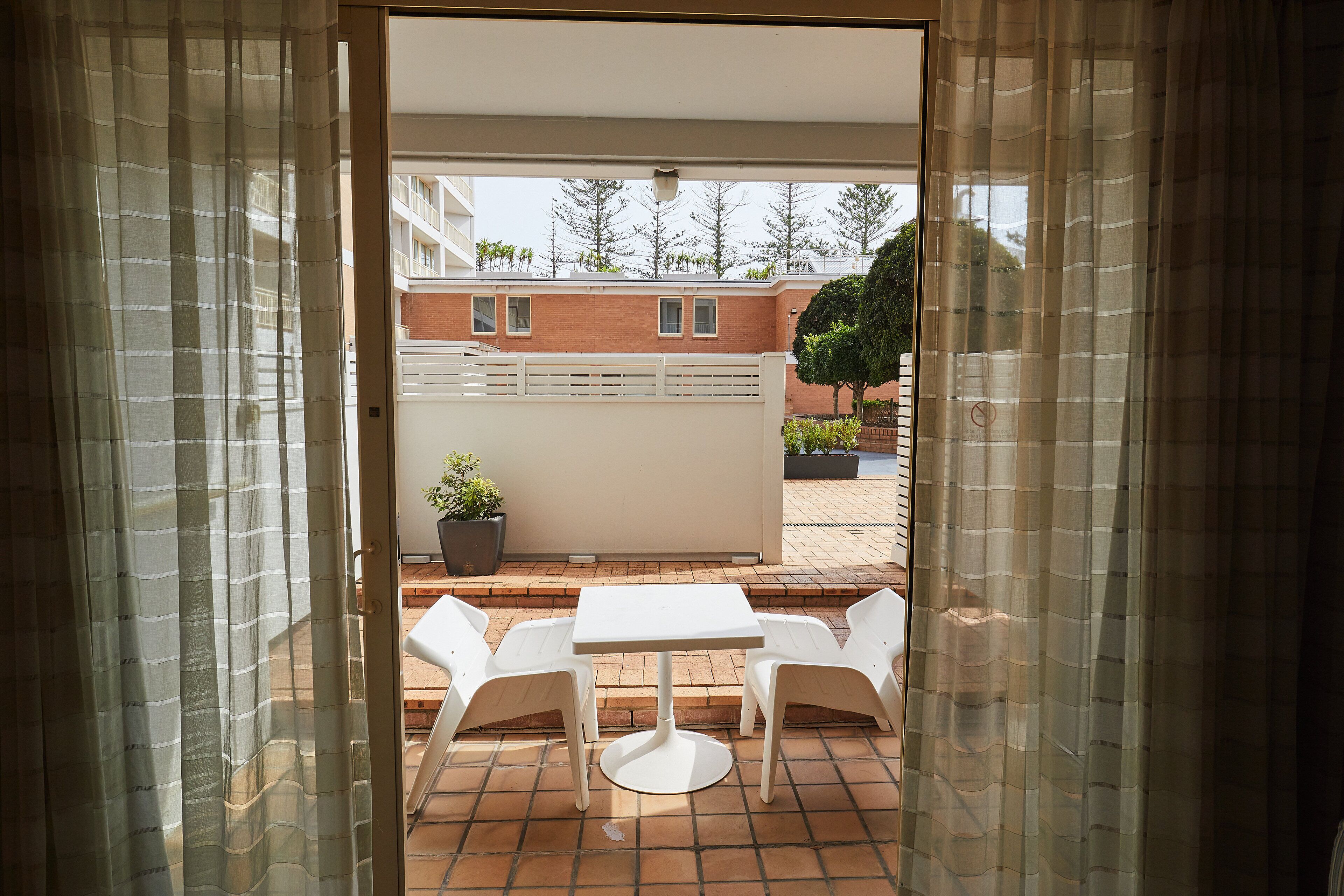Une porte ouverte donne sur un patio où se trouvent une table blanche et deux chaises, un pot de fleurs et un mur de briques percé de fenêtres.