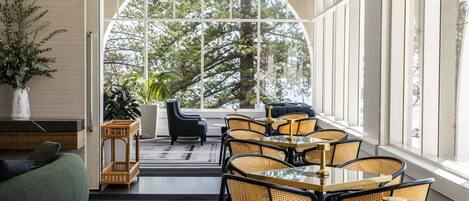 A modern dining area with a glass table and wicker chairs, large windows with a view of trees, and a green sofa.