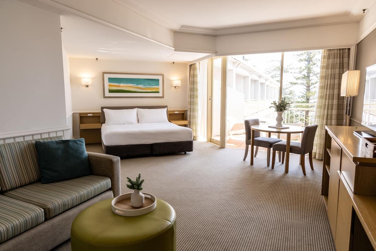 Una habitación de hotel con una cama, un sofá, una mesa de comedor con sillas, una mesita con una planta y un escritorio con una lámpara.