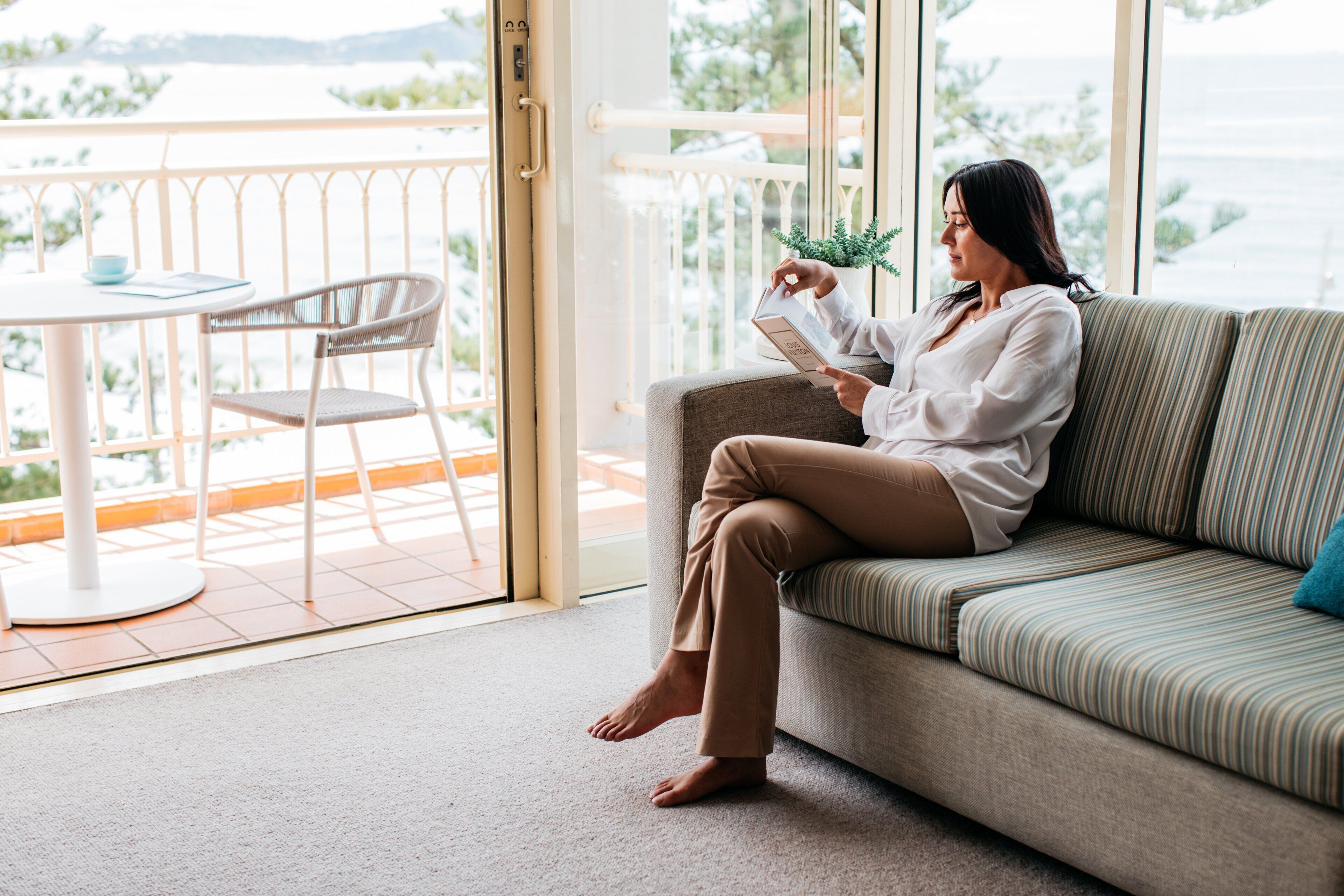 A woman sitting on a sofa reading a book in a room with a balcony, a table, and a chair.