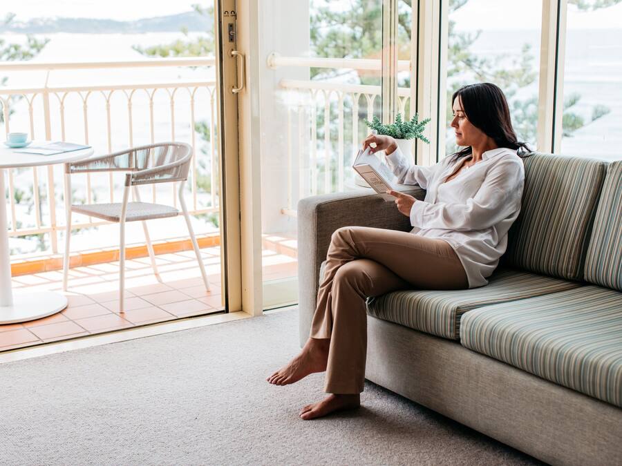 A woman sitting on a sofa reading a book in a room with a balcony, a table, and a chair.