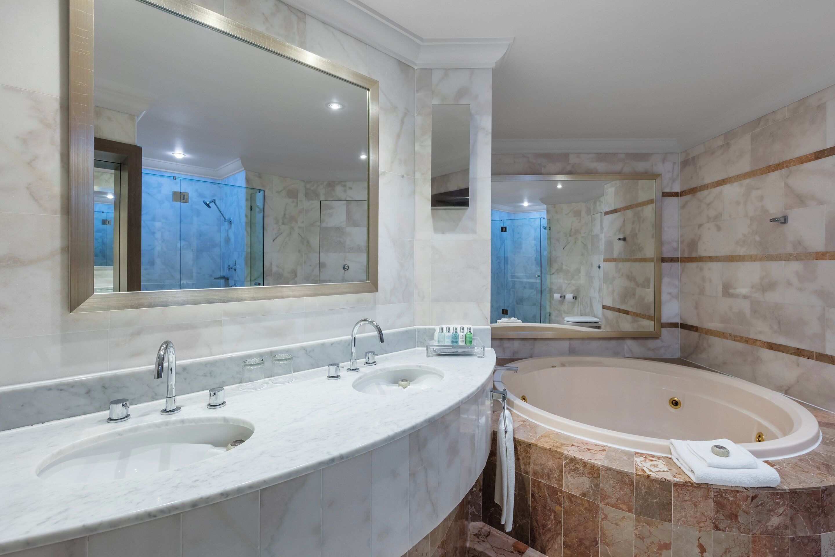 A modern bathroom with a large mirror, two sinks, a bathtub, and marble tiles.