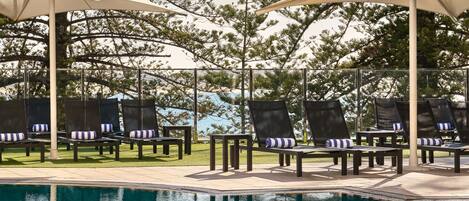Un área junto a la piscina con sillas de descanso y sombrillas, rodeada de árboles.