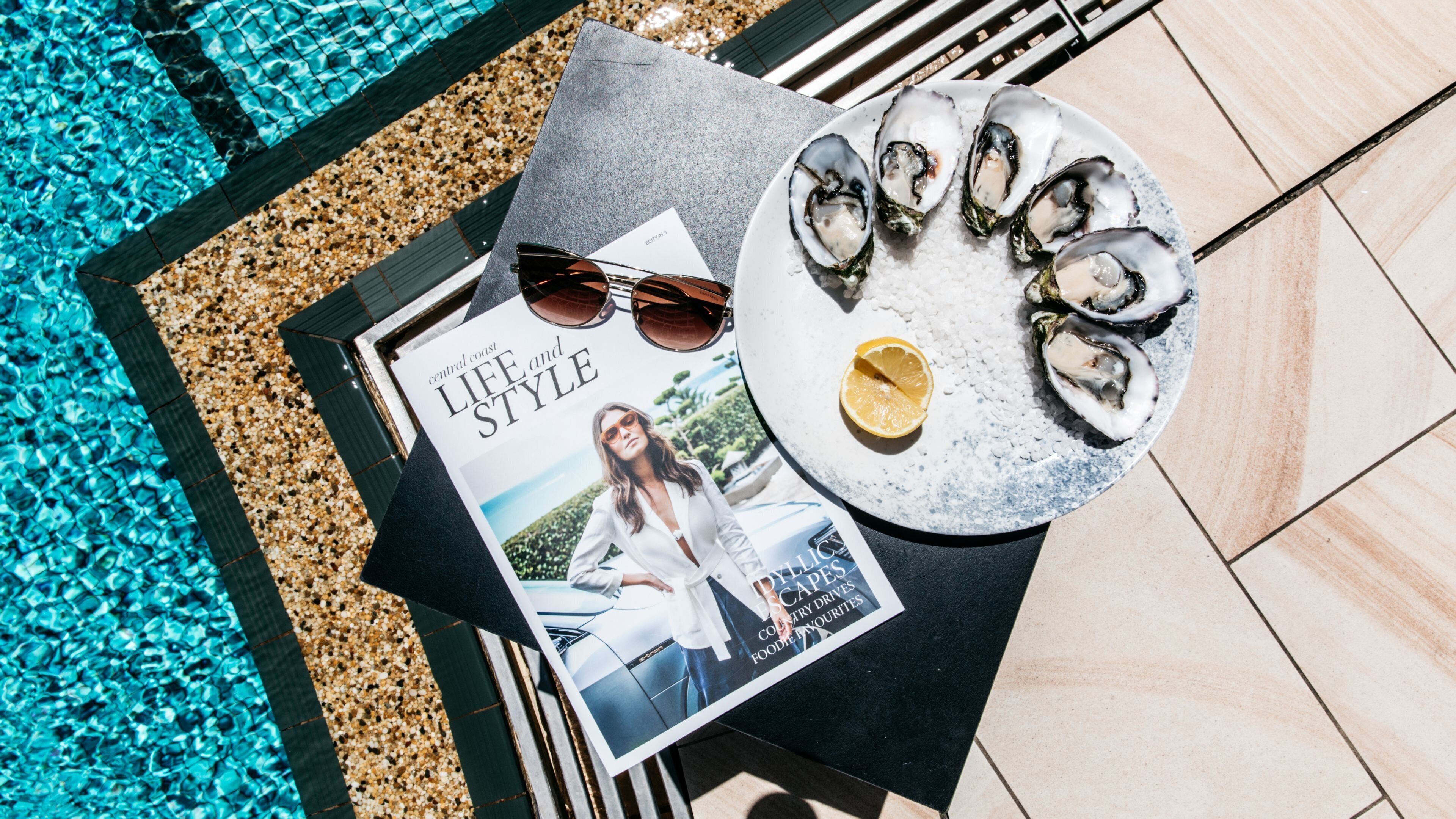 A table with a magazine, sunglasses, a plate of oysters, and a lemon wedge.