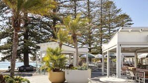 An outdoor seating area with round tables and chairs, surrounded by large planters with various plants, overlooking a body of water. - Crowne Plaza Terrigal Pacific by IHG (Terrigal)