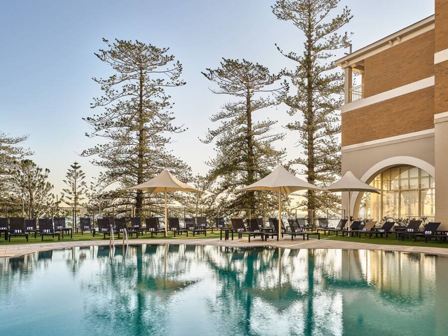 Un espace piscine doté de chaises longues et de parasols, entouré de grands arbres et bordé d’un bâtiment aux larges fenêtres.