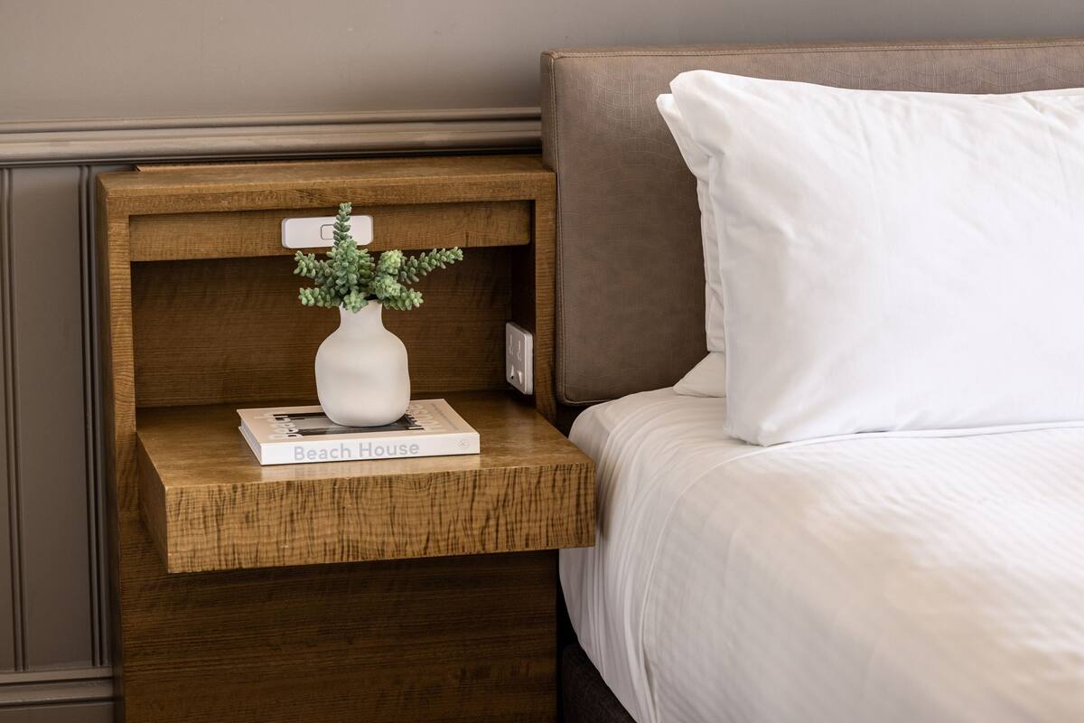 Una cama bien hecha con un almohadón blanco y una mesita de noche de madera. Sobre la mesita, hay un florero blanco con follaje verde y un libro titulado