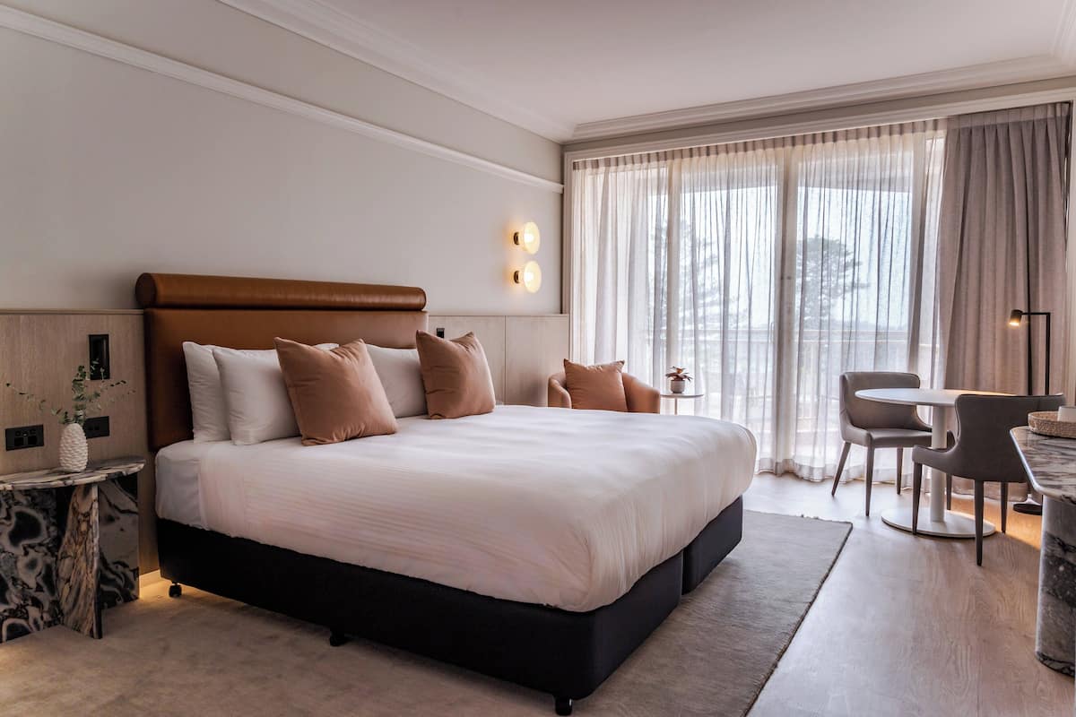 Habitación de hotel moderna con una cama grande, una pequeña mesa con sillas y vistas al exterior a través de cortinas traslúcidas.