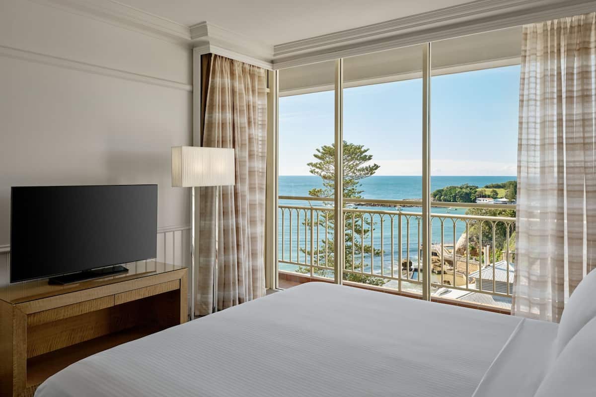 Habitación de hotel con una cama grande, un televisor de pantalla plana sobre un mueble de madera, un balcón con vistas al mar y follaje.