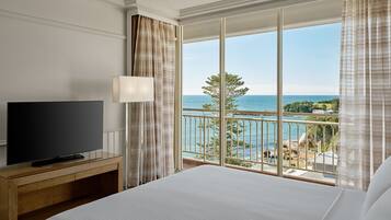 Habitación de hotel con una cama grande, un televisor de pantalla plana sobre un mueble de madera, un balcón con vistas al mar y follaje.