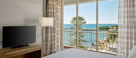 A hotel room with a large bed, a flat-screen TV on a wooden console, a balcony with a view of the sea and greenery.