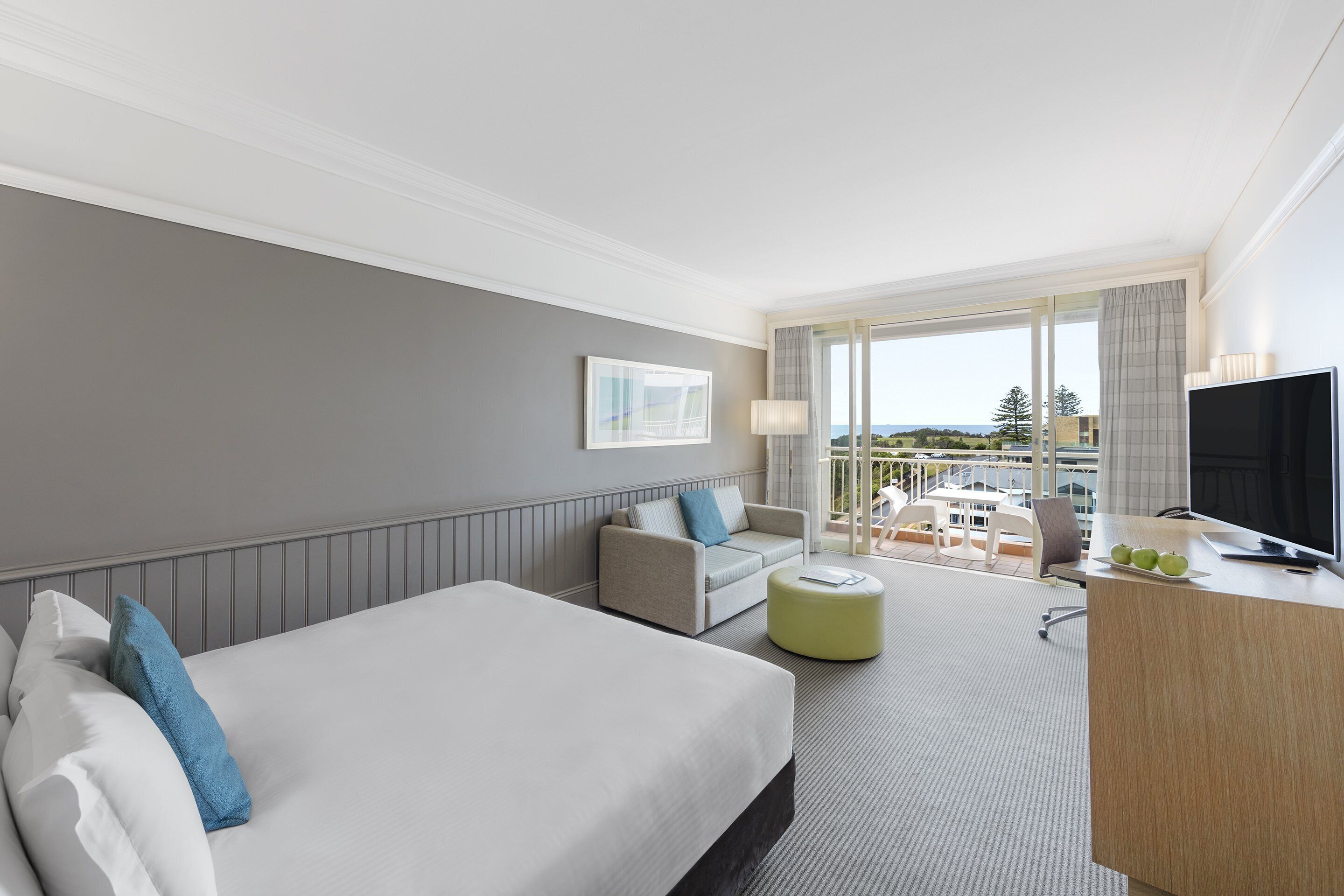 Habitación de hotel con una cama grande, un sofá, un escritorio con televisor y un balcón con vistas.