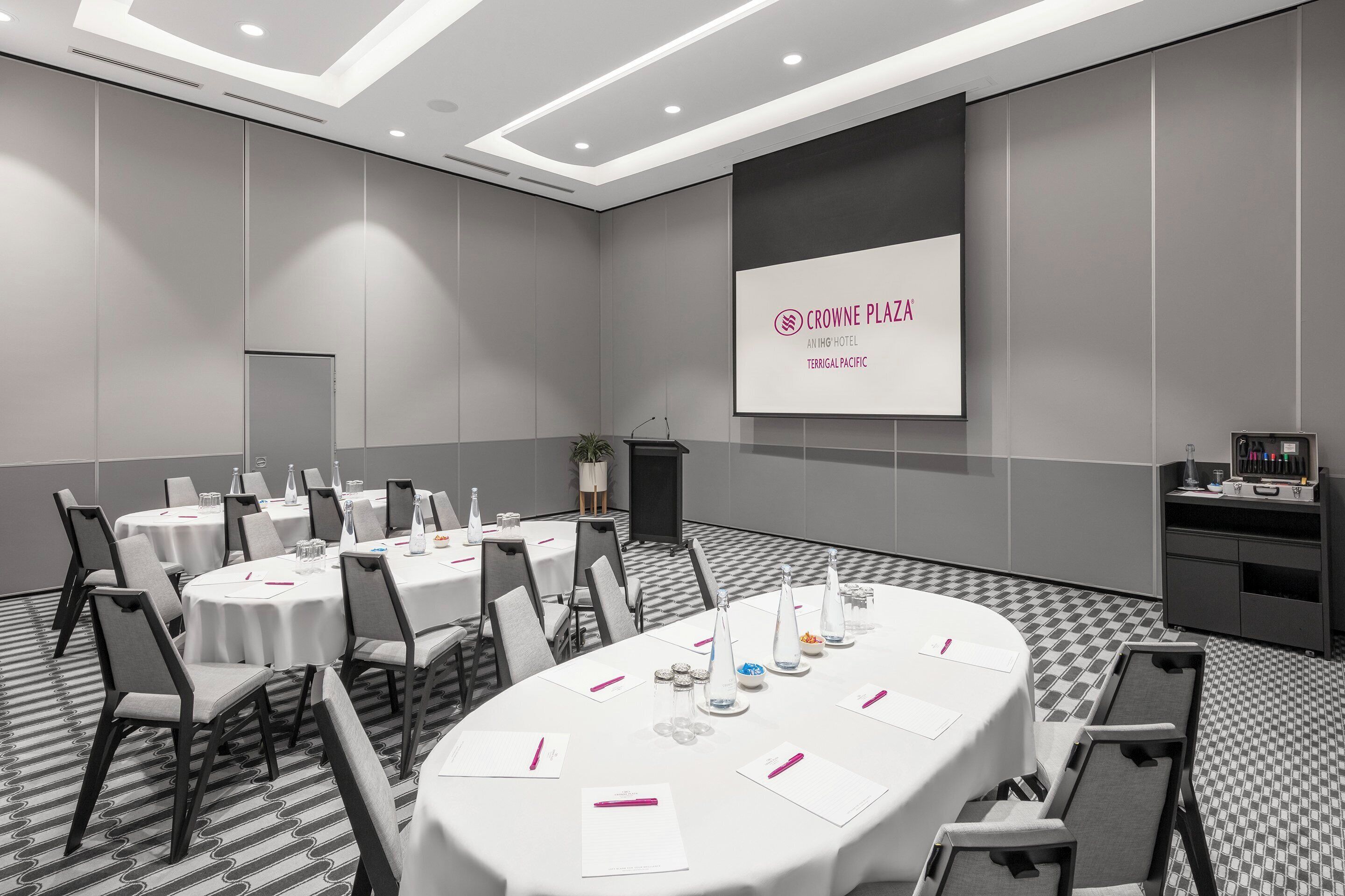 Un salón de conferencias con mesas redondas, sillas y una pantalla de presentación que muestra 'CROWNE PLAZA AN IHG HOTEL TERRIGAL PACIFIC'.