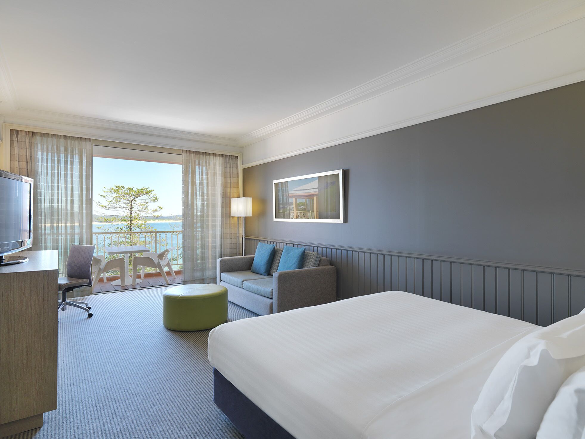 A hotel room with a bed, a desk with a TV, a sofa, a chair, and a balcony with a view of the sea.