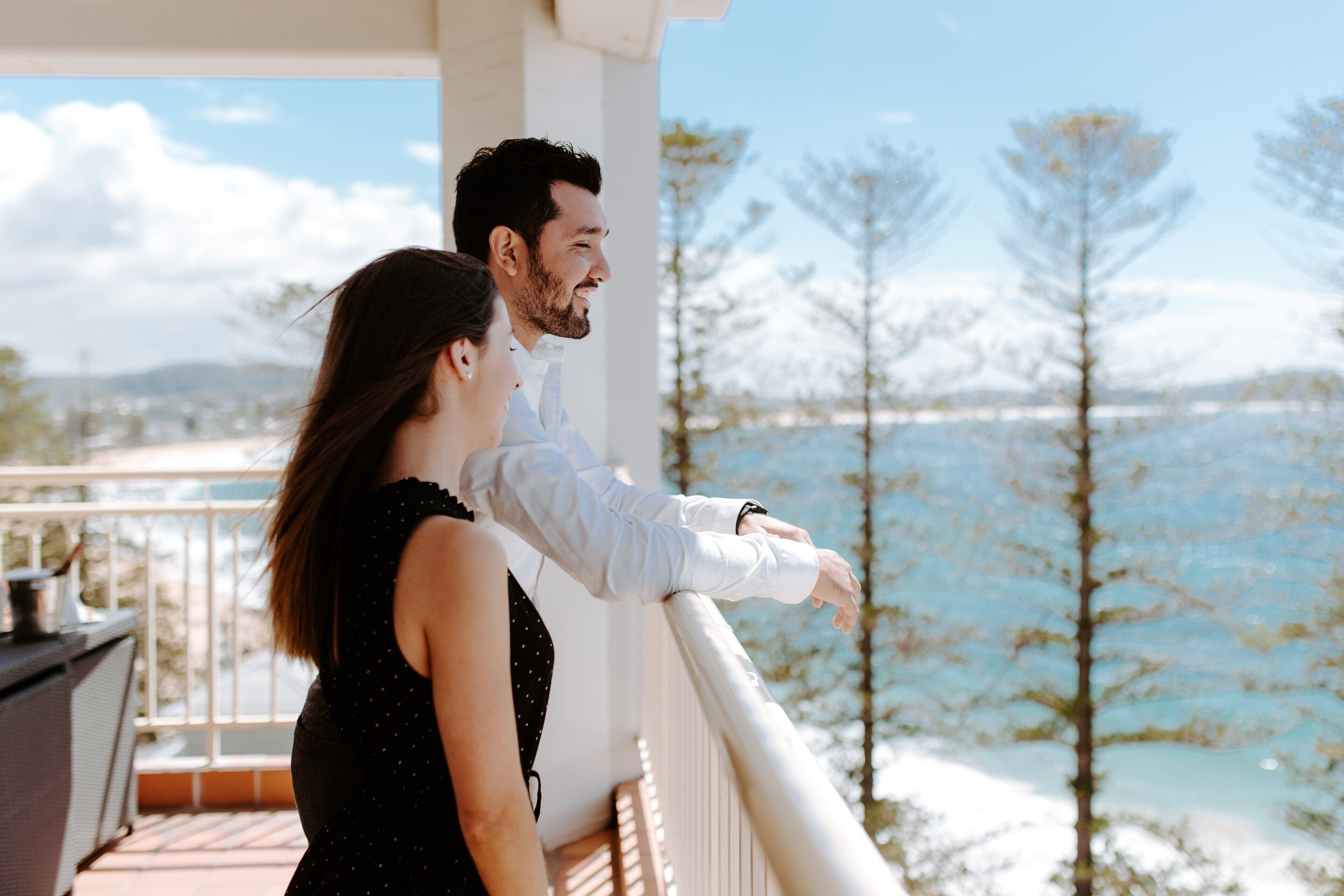 Un couple se tient sur un balcon, d’où l’on peut admirer un paysage magnifique composé d’arbres et d’eau.