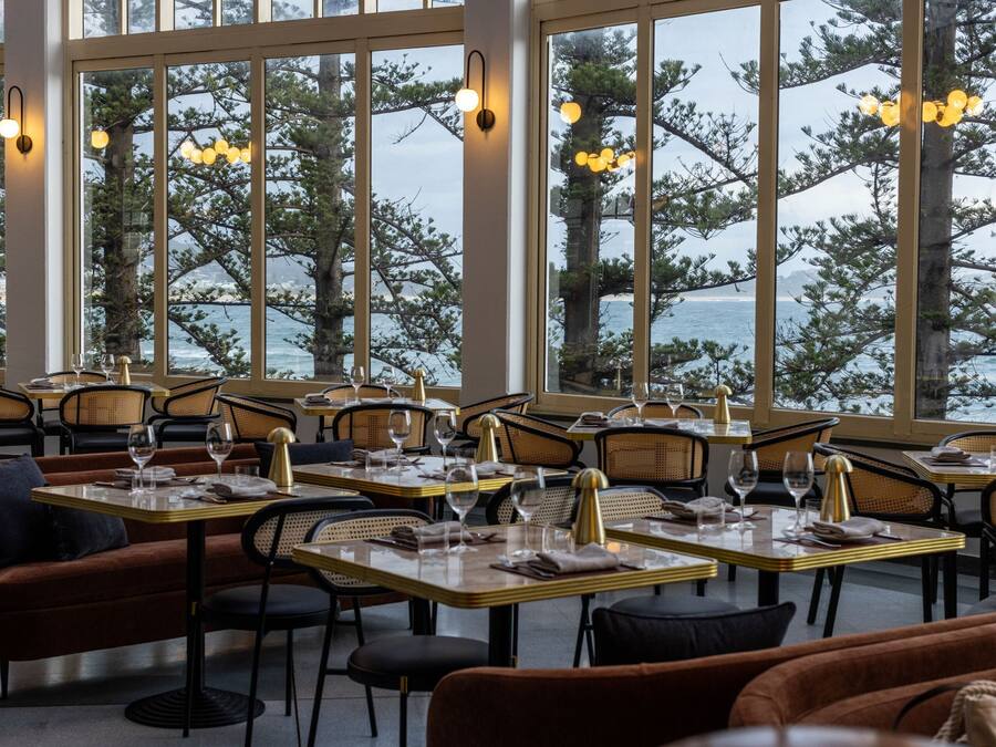 A restaurant with tables set for dining, large windows with a view of trees and water, and warm lighting.