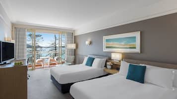 A hotel room with two beds, a TV, a desk, and a balcony with a view of trees and mountains.