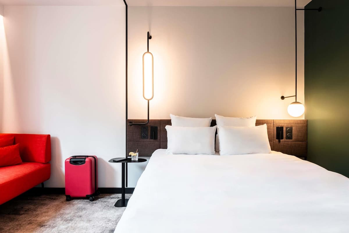 A hotel room with a bed, a red armchair, a suitcase, and two bedside tables with lamps.