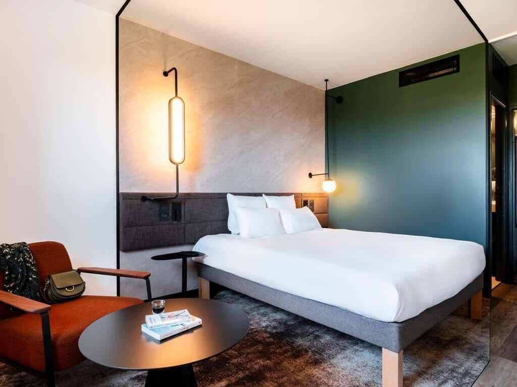 A modern hotel room with a bed, a round table, a chair, and a wall-mounted light fixture.