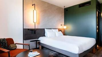 A modern hotel room with a bed, a round table, a chair, and a wall-mounted light fixture.
