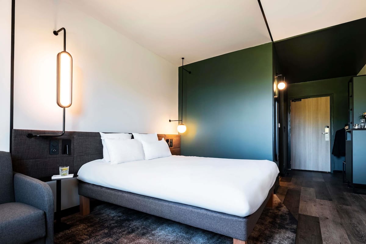 A modern hotel room with a large bed, a bedside table, a chair, and a wall-mounted lamp.