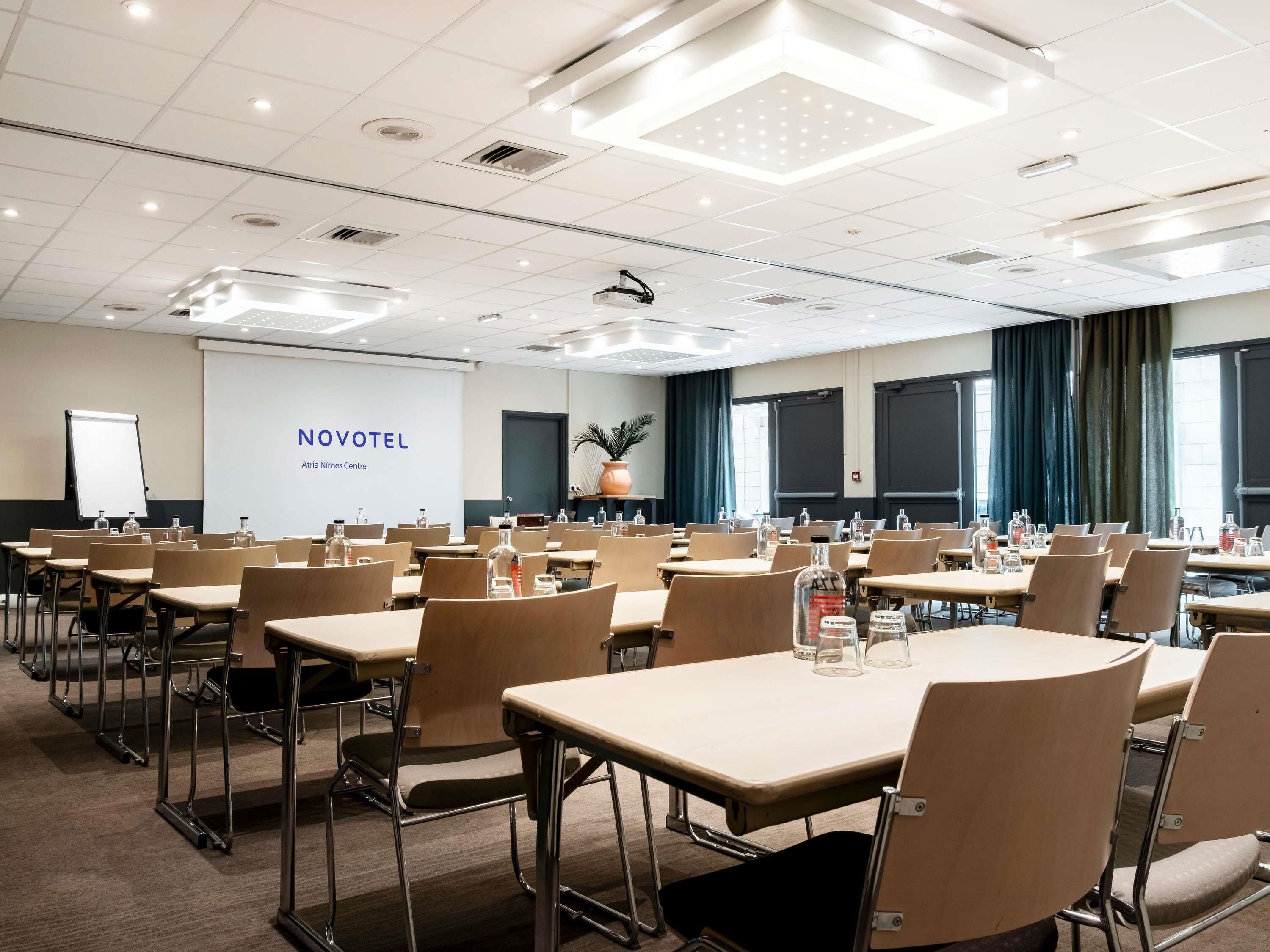 Een vergaderruimte met tafels en stoelen, een beamer en een scherm waarop 'NOVOTEL Atrium Nimes Centre' staat.