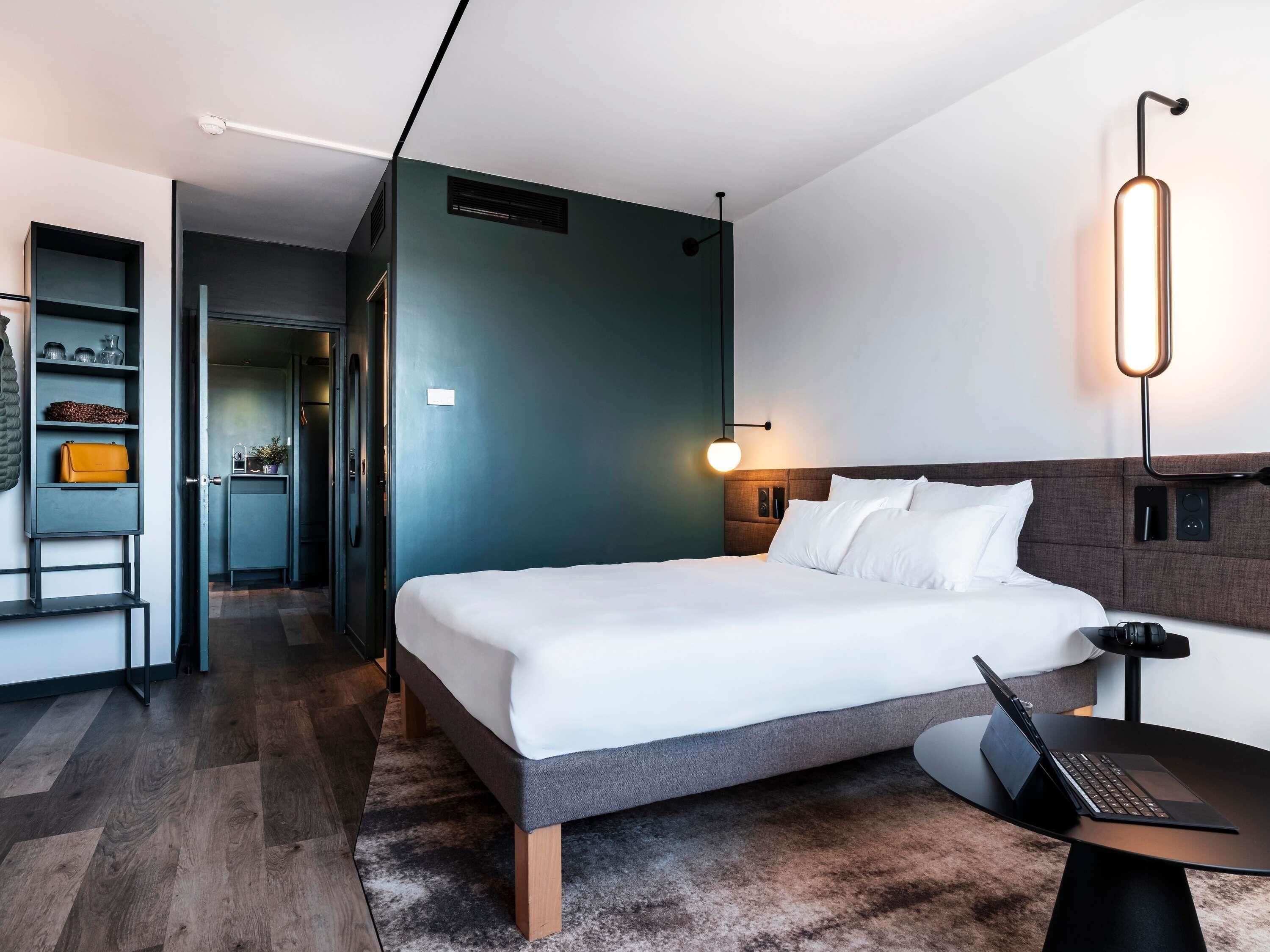 A modern hotel room with a large bed, a desk with a laptop, a bedside table, and a wall-mounted lamp.