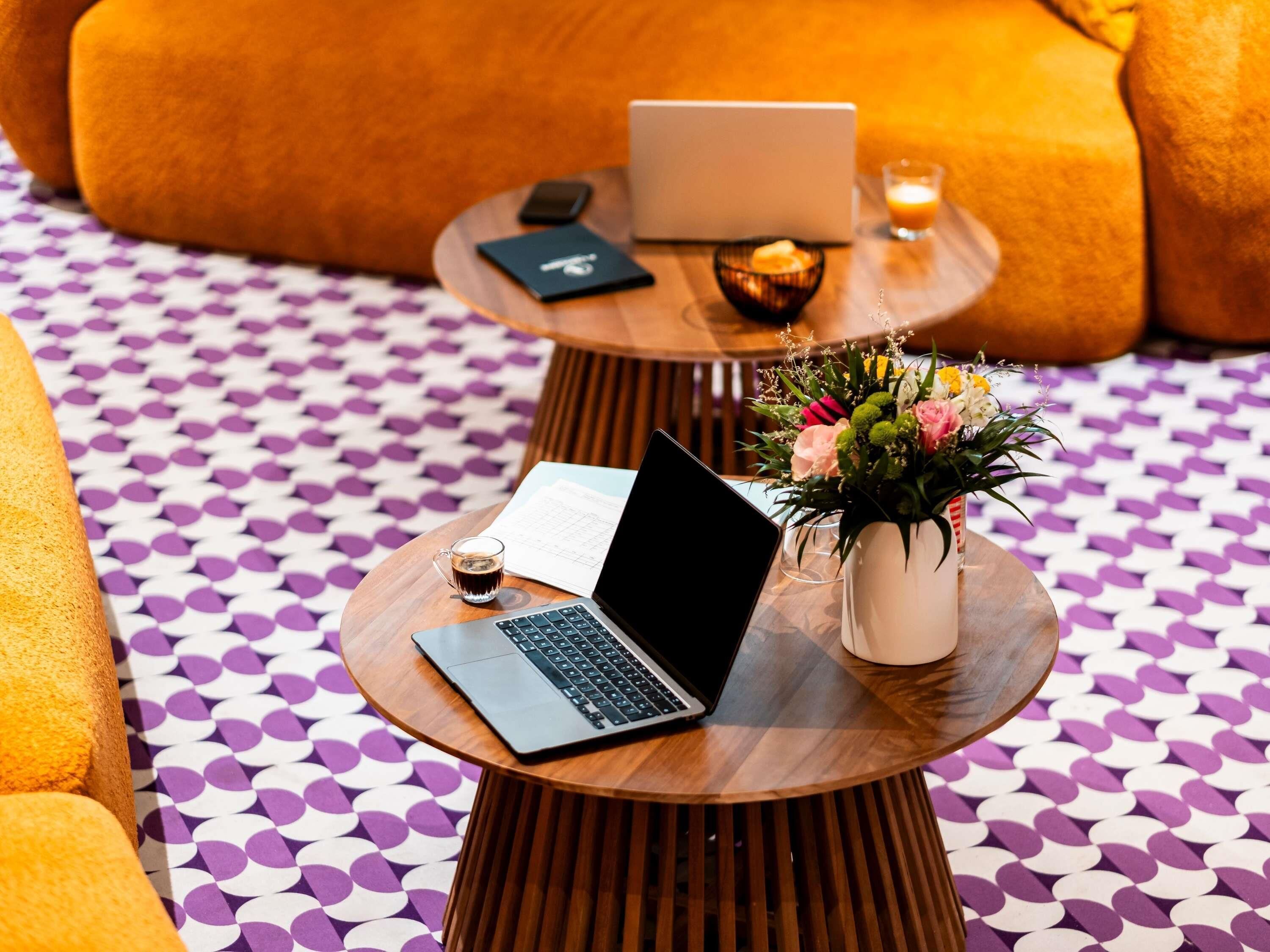 A round wooden table with a laptop, a glass of coffee, a smartphone, a candle, and a vase with flowers.