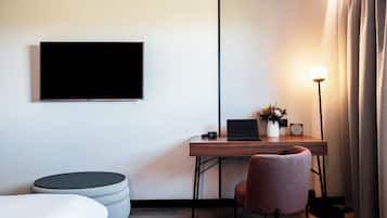 Une chambre d’hôtel moderne équipée d’un téléviseur à écran plat fixé au mur, d’un bureau en bois avec un ordinateur portable, d’une petite plante en pot et d’une lampe sur pied.