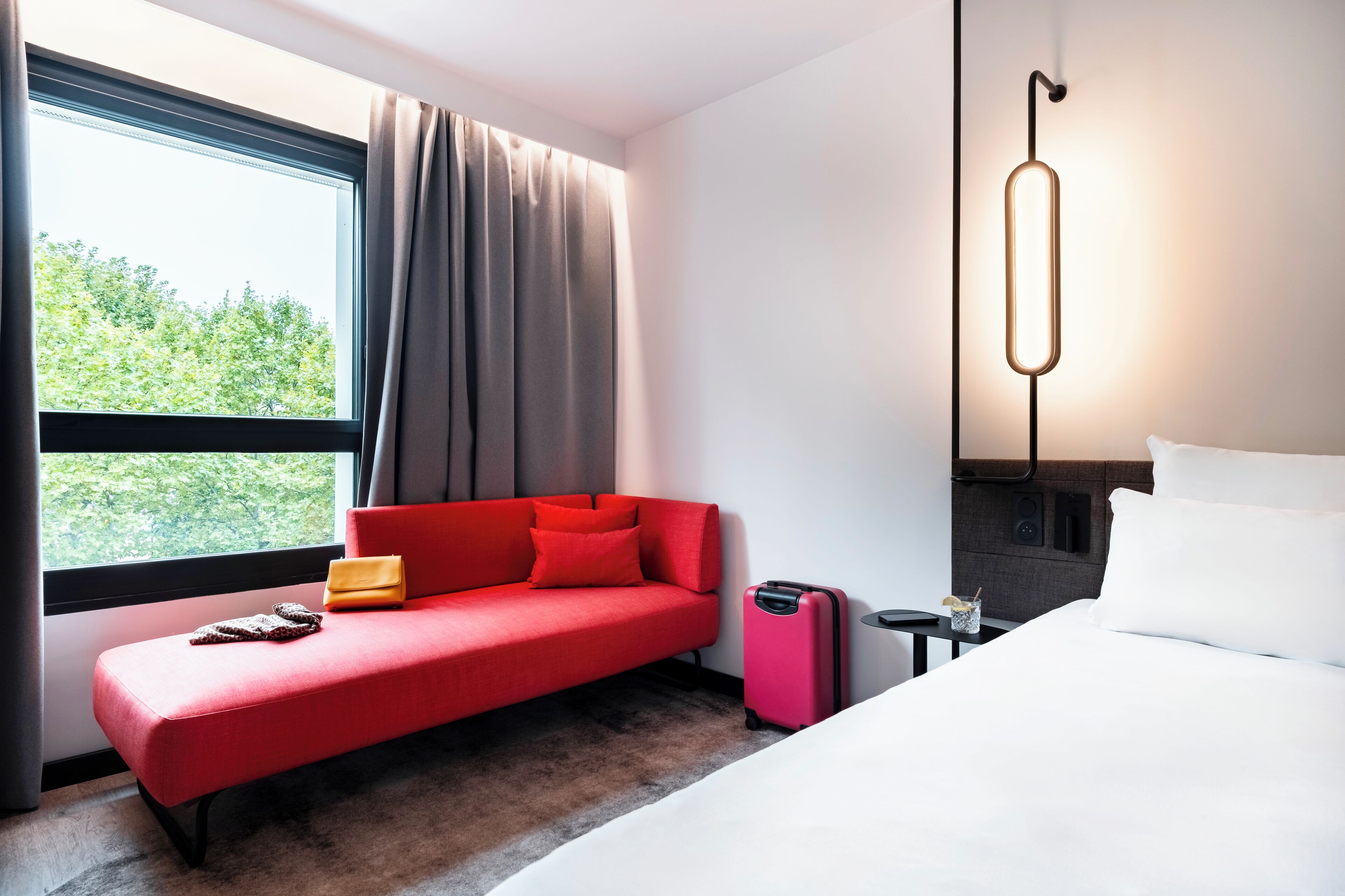 A modern hotel room with a red sofa, a white bed, and a large window with curtains.