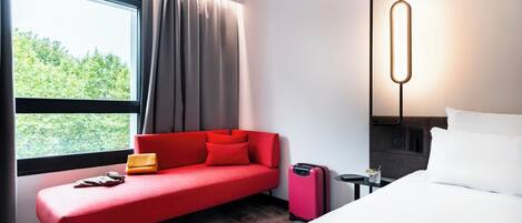 A modern hotel room with a red sofa, a white bed, and a large window with curtains.
