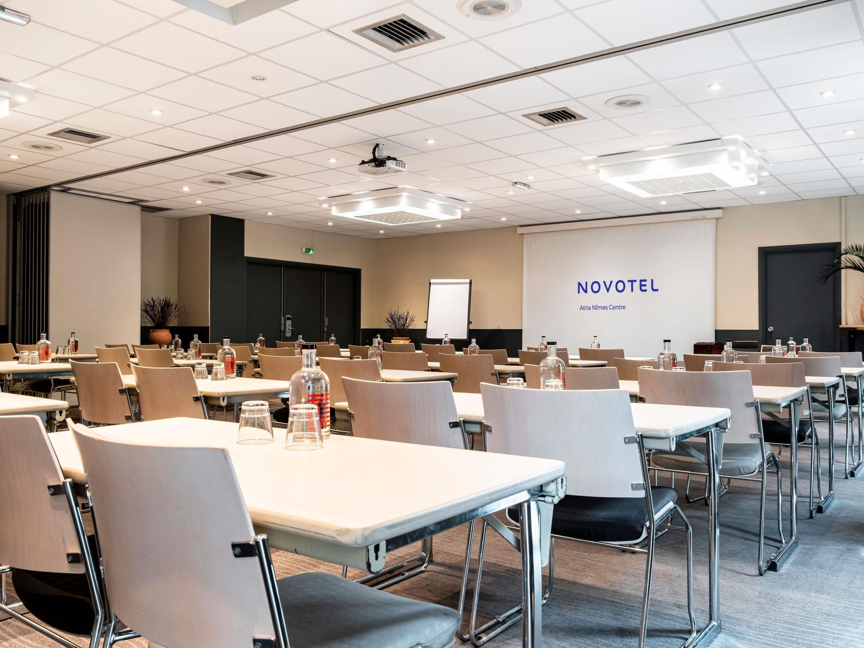 A conference room with tables, chairs, and a projection screen displaying 'NOVOTEL Atria Nimes Centre'.