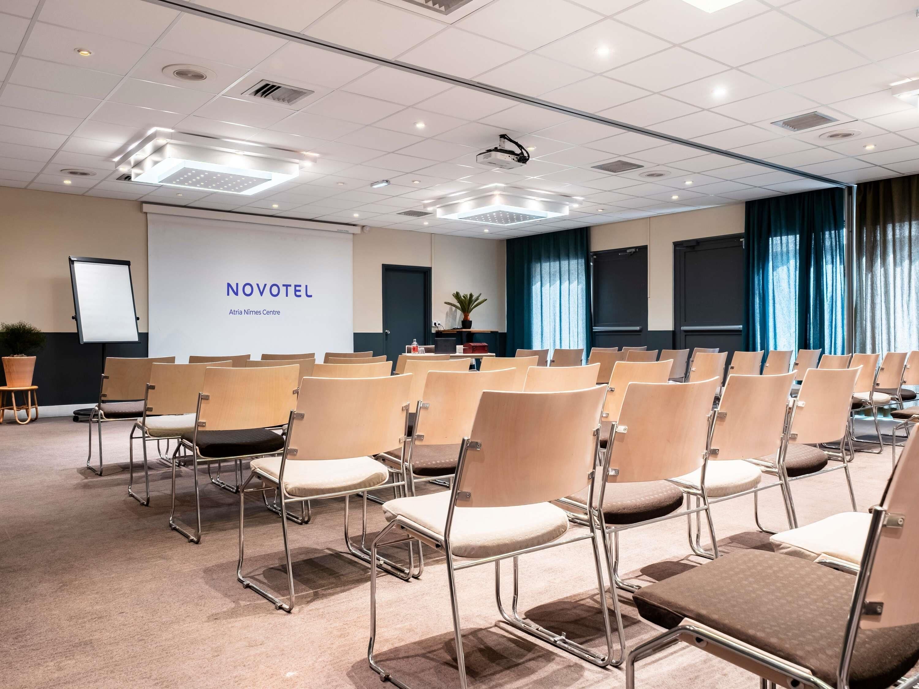 A conference room with rows of chairs, a projector, and a screen displaying 'NOVOTEL Atria Nimes Centre'.