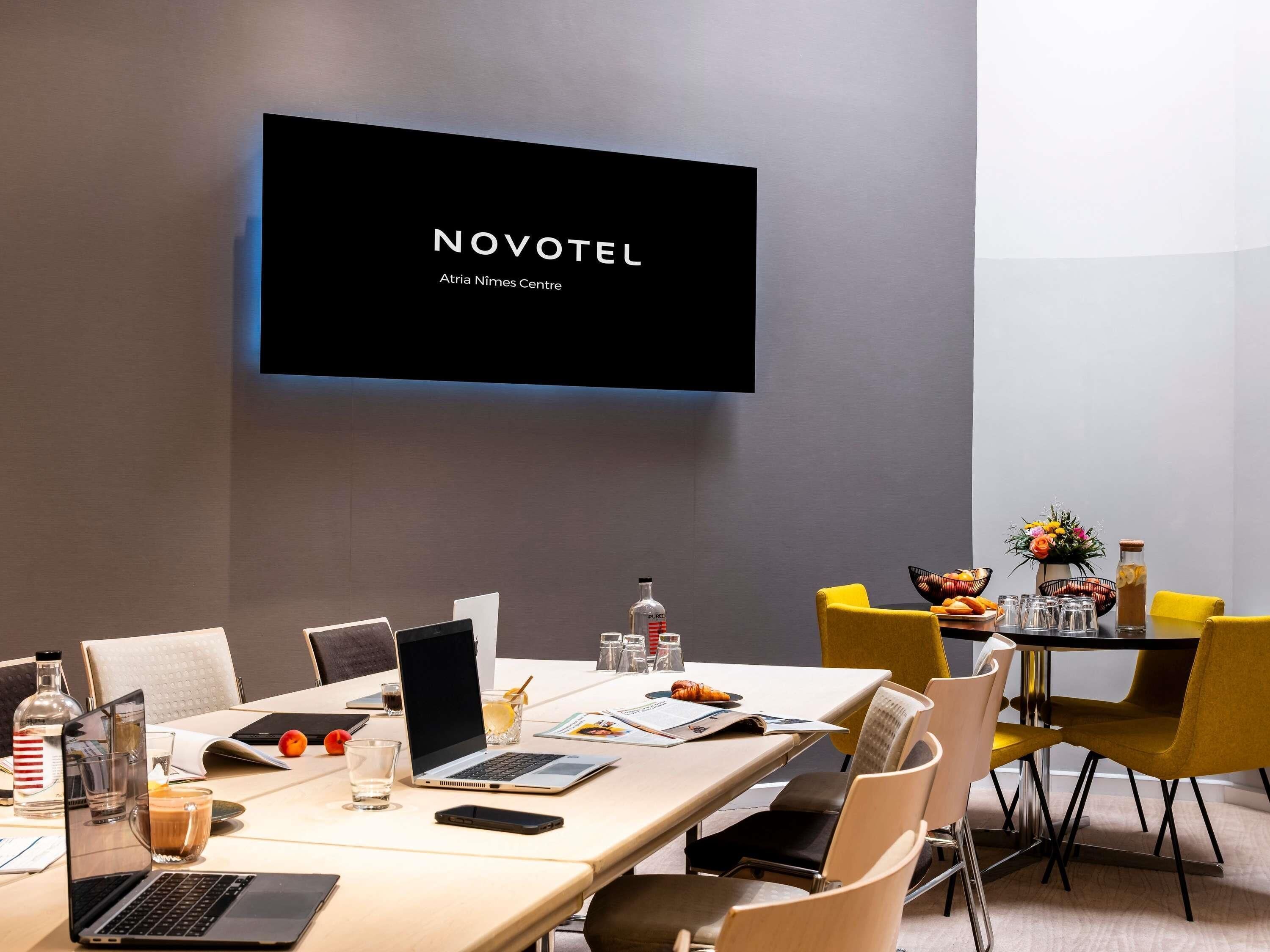 A modern hotel conference room with a flat-screen TV displaying 'NOVOTEL Atria Nimes Centre,' a long table with laptops, chairs, and a small table with food and drinks.